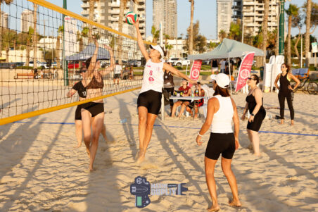 צילום ספורט של אליפות ישראל בכדורשת בחוף הים אשדוד, שחקנית מזנקת להנחתה מעל הרשת בזמן משחק מותח מול קהל ומתחרות, תיעוד של רגע השיא